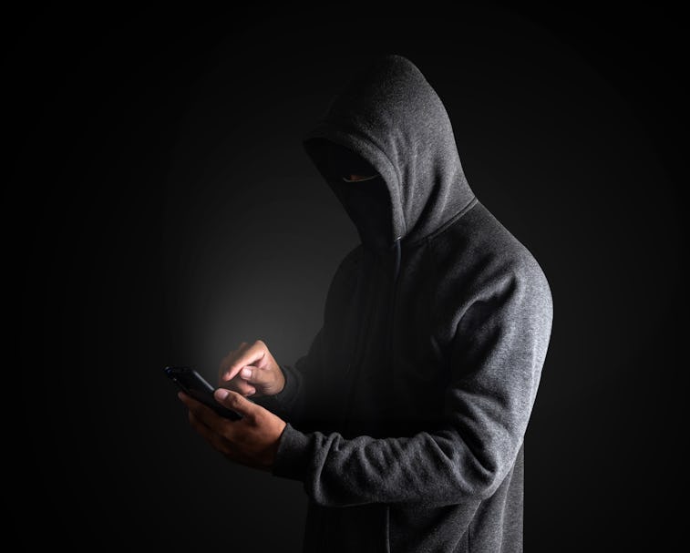 Hacker Using Smartphone. Men in black clothes with hidden face looks at smartphone screen on black b...