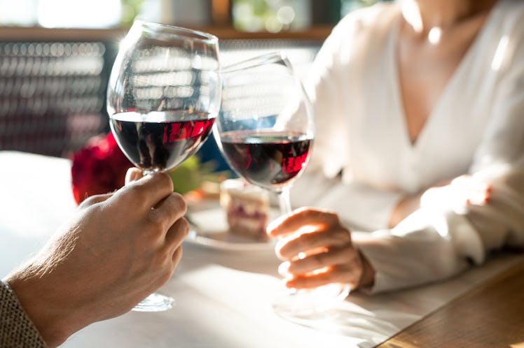 Hands of young man and his girlfriend clinking with wineglasses over table while enjoying romantic d...
