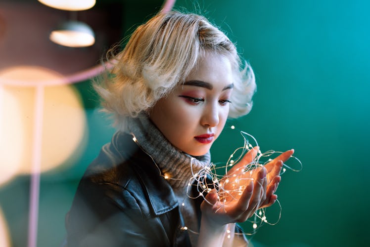 Young adult blonde female asian holding string of fairy lights with bokeh around, selective focus