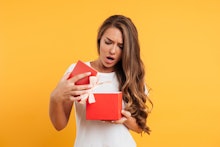 Portrait of an upset disappointed girl opening gift box isolated over yellow background