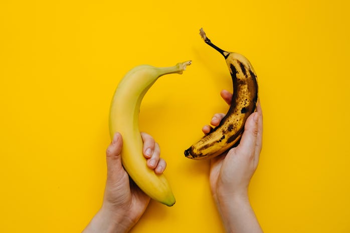 Human hands holding two bananas: fresh and ripe, on bright yellow background.