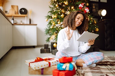 A young woman on the background of a Christmas tree with gifts, with a tablet has a video call or vi...