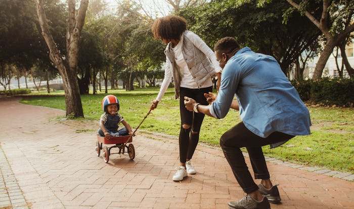 Boy sitting in a small wagon pulled by his mother, while father standing with his arms outstretched....