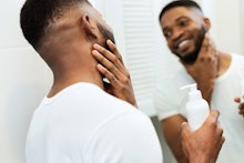 Facial care. Young black man applying moisturizer on beard in bathroom, looking in mirror