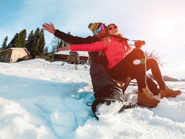 A happy couple slides down a snowy hill on a vintage sled while the sun shines on them.