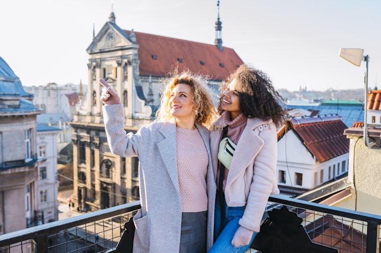 Two happy friends in coats smile on a rooftop and enjoy the views on a sunny winter morning.