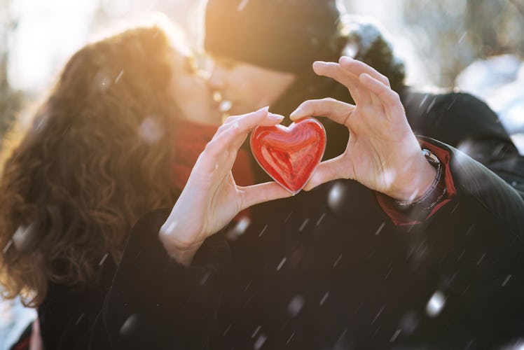 A couple kisses and holds out a plastic heart in front of them while it snows.