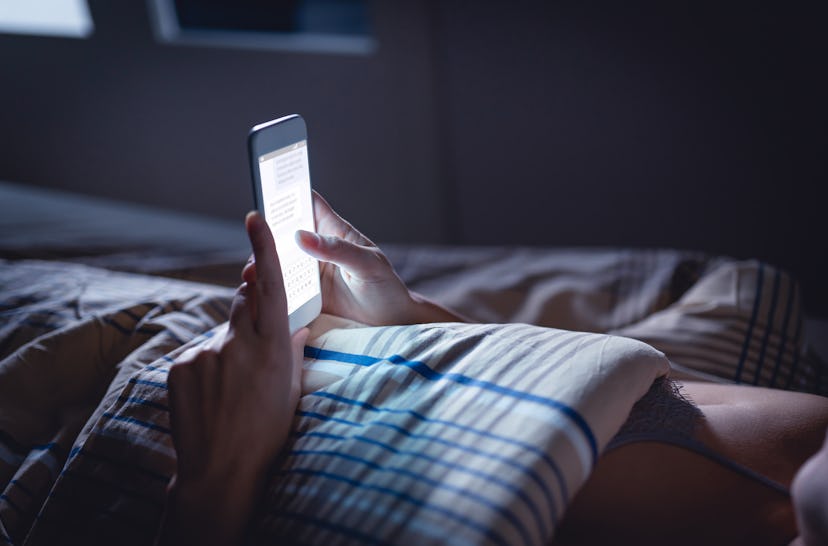 A woman using her smartphone at night while lying in bed, and covered with a blanket