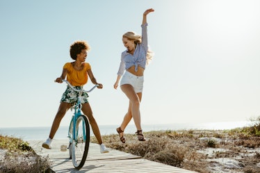 Two girls laugh while one rides a bike and the other dances down the boardwalk on a sunny day.