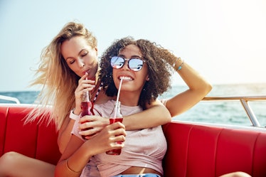 Two friends sip on soda while riding on a boat with red seats, and laughing on vacation. 