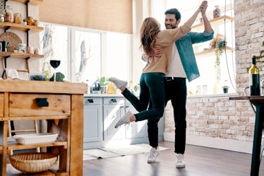 A couple dressed in casual outfits and sneakers laugh and embrace in the kitchen on a sunny day.