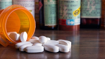 White prescription pills spilled onto a table with many prescription bottles in the background. Conc...