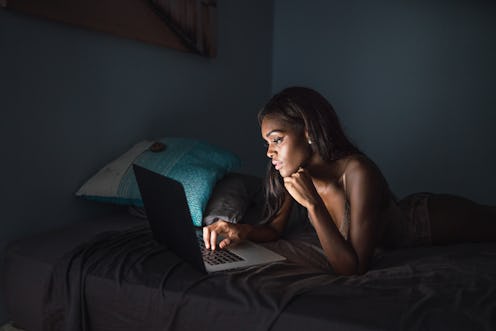 Beautiful African American woman lying on bed in dark bedroom and surfing laptop