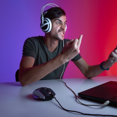Portrait of an angry gamer playing video games on computer in dark room wearing headphones and showi...