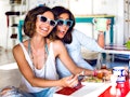 Two friends wearing sunglasses smile while enjoying brunch together at a colorful, bright restaurant...