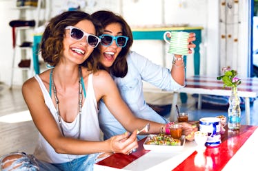 Two friends wearing sunglasses smile while enjoying brunch together at a colorful, bright restaurant...