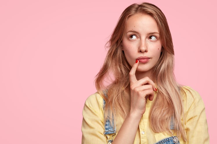 Thoughtful female youngster keeps fore finger near lips, being deep in thoughts, thinks about coming...