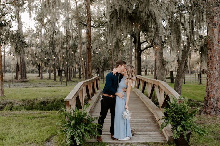 A bridesmaid in her blue dress kisses her date at a wedding while standing on a bridge under string ...