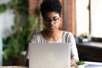 Focused african American millennial girl in glasses sit at desk working at laptop browsing internet,...