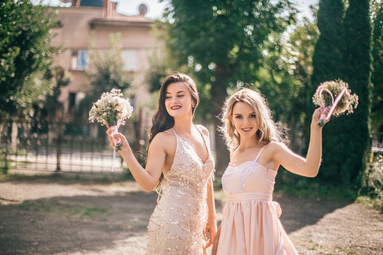 Two bridesmaids dressed in pink dresses hold up their bouquets at a wedding they're attending. 