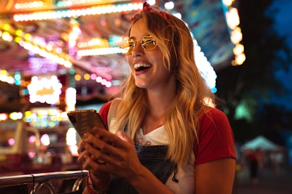 A blonde woman wearing sunglasses and a red headband laughs while holding her phone at a carnival at...