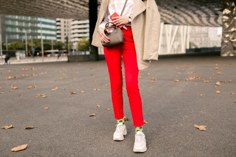 Street fashion autumn details of young progressive hipster business woman, red pants, funny socks and trendy ugly sneakers, beige classic coat, modern buildings around, phone and accessories.