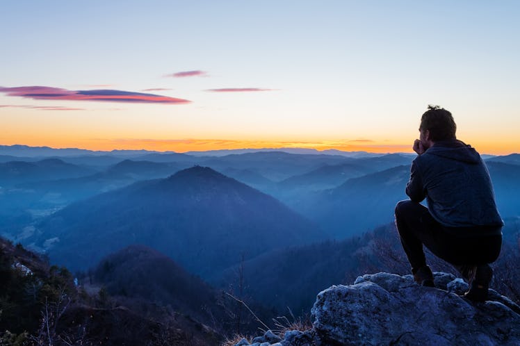 Male hiker crouching on top of the hill and enjoying scenic view of twilight landscape below. Hiking...