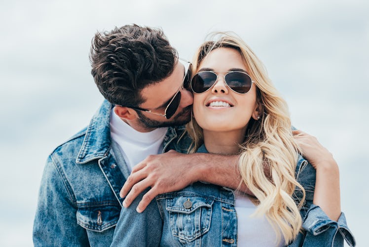 A man kisses his girlfriends while wearing denim jackets and sunglasses on a sunny day.