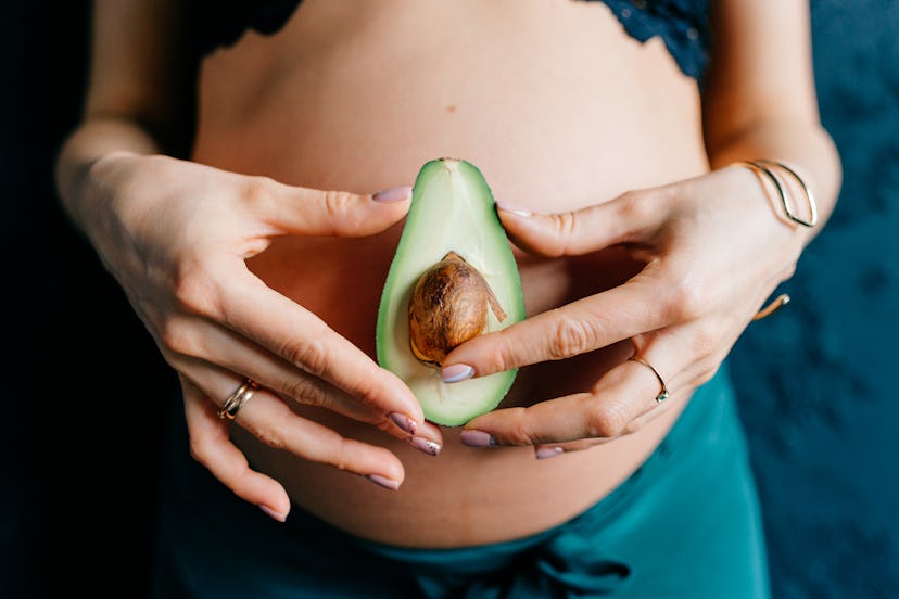 Young girl holding one half avocado on background of her pregnant belly. expectant mother is waiting...