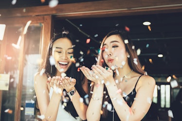 Two women in dresses blow confetti in the air at a restaurant while celebrating a birthday.