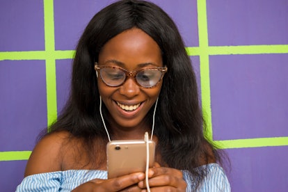 A happy woman standing in front of a bright purple wall looks down at her phone.