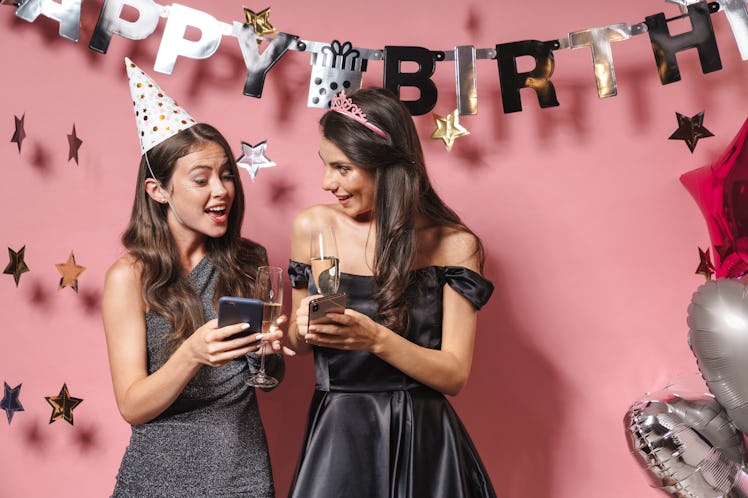 Two friends dressed up for a birthday celebration hold champagne flutes and their phones.