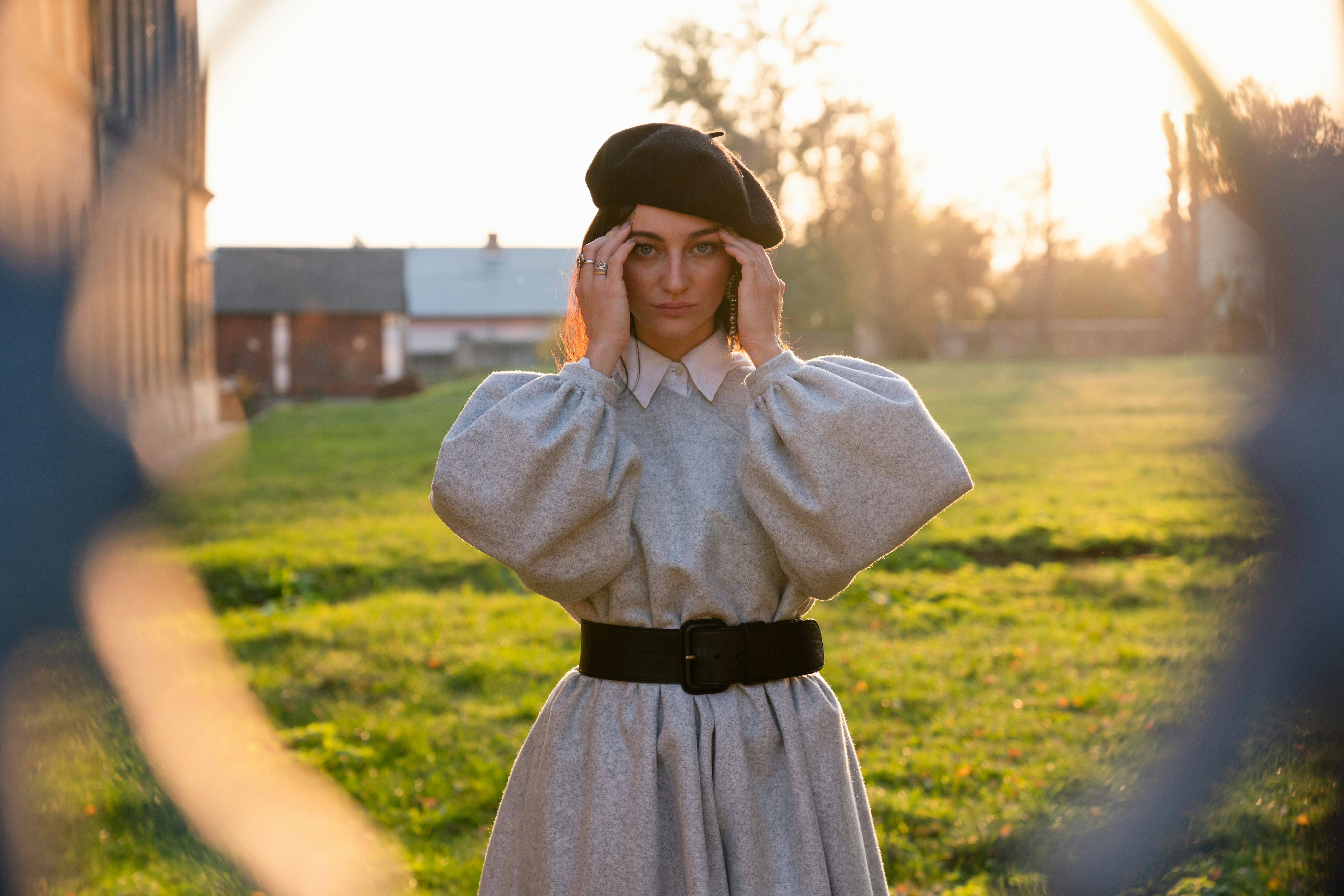 Sad young woman in black beret and grey dress holds her hands near her head, have a headache, stands...
