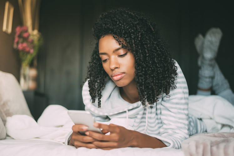 Beautiful serious thoughtful and sad black woman with curly hair using smartphone on bed