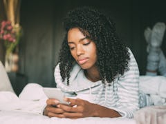 Beautiful serious thoughtful and sad black woman with curly hair using smartphone on bed
