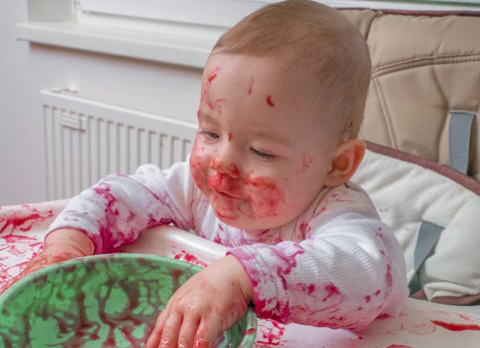 Messy dirty little baby is eating with hands from bowl.