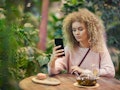 Portrait of young beautiful woman with curly hair using her phone while drinking tea in green cafe