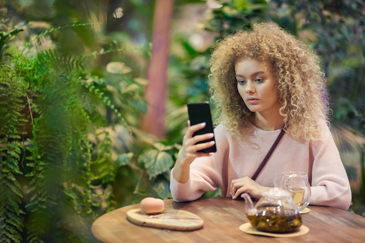 Portrait of young beautiful woman with curly hair using her phone while drinking tea in green cafe