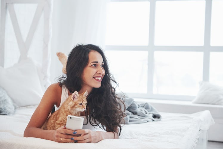 A woman is typing on her phone next to her orange kitten, while laying in bed. 