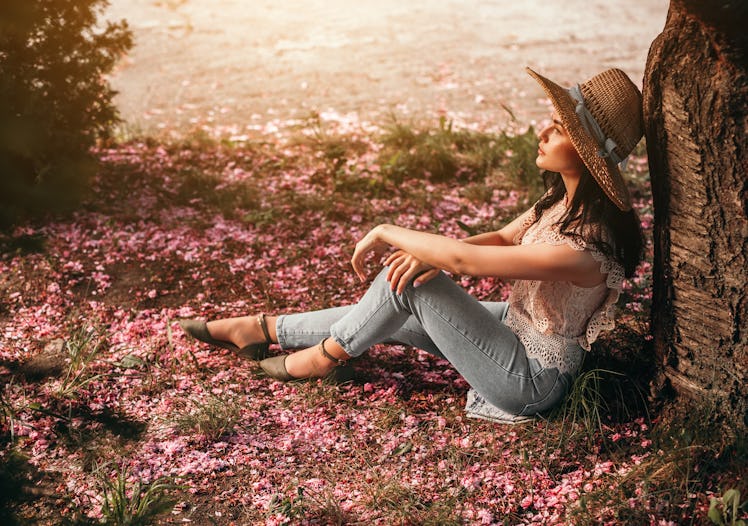 Side view of stylish lady sitting on ground covered with pink flower petals and leaning on tree trun...