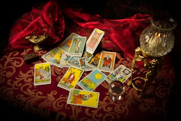 A pile of tarot cards lie scattered and spread across a table top surrounded by multiple occult item...