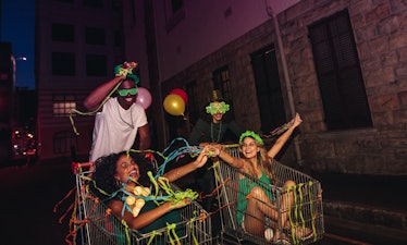 A group of four friends has fun in shopping carts at night with confetti and balloons, celebrating S...