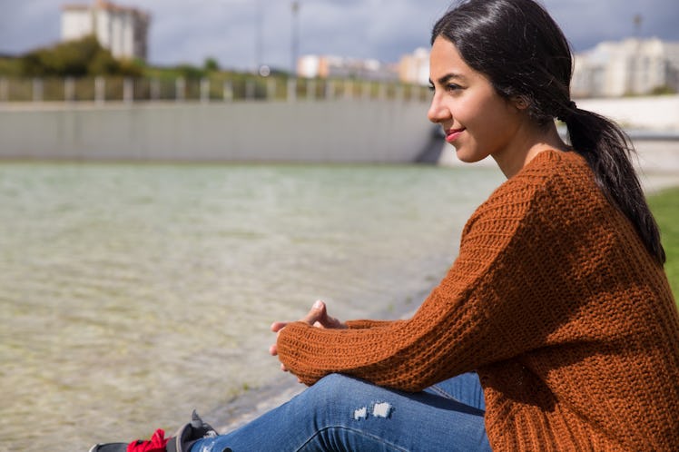 Dreamy pretty woman sitting by city river. Young lady looking away and sitting with distant building...