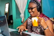happy mature african woman is sitting at home working laptop, listening to music, and drinking coffe...