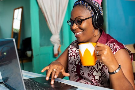 happy mature african woman is sitting at home working laptop, listening to music, and drinking coffe...