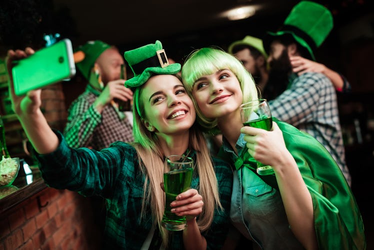 Two girls dressed up for St. Patrick's Day at a bar smile with their green beers for a selfie.