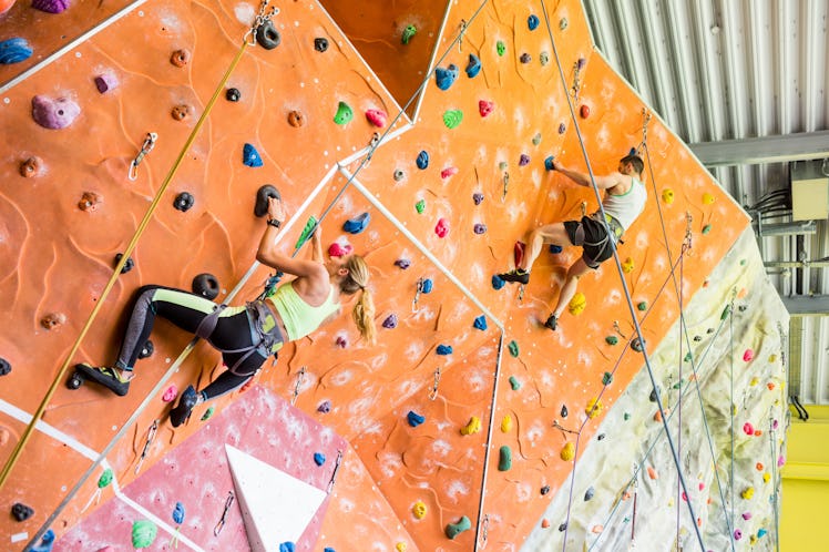 An athletic couple rock climbs on a colorful wall in a gym.