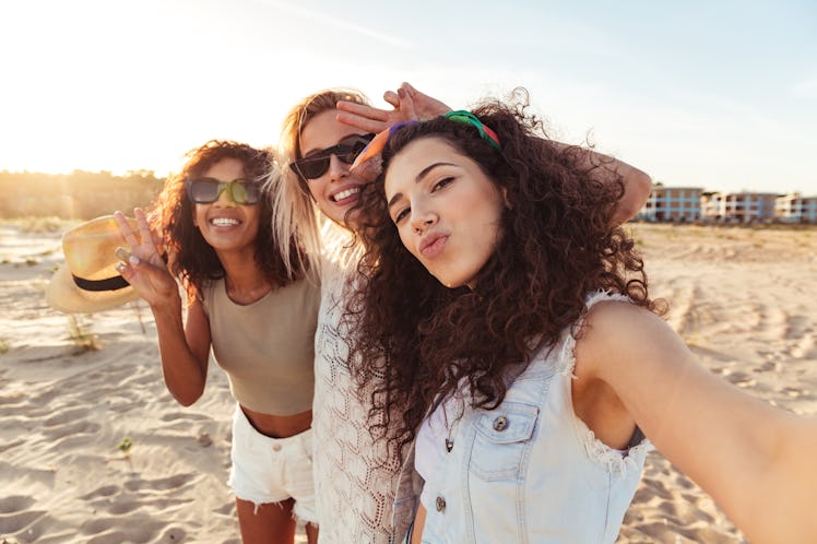 Three happy friends dressed in summery outfits smile for a selfie on a beach, which they'll use as a...