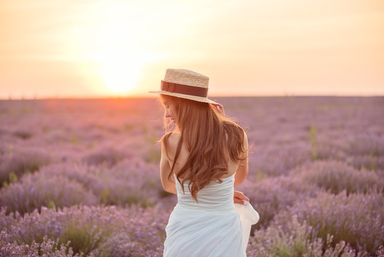 Back view of beautiful girl in a white dress and hat in a lavender field at sunset