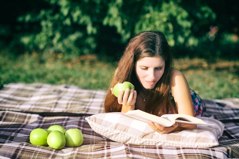 Cute young girl lying on a pillow, eating green apple and read a book in a park. A perfect picnic ph...